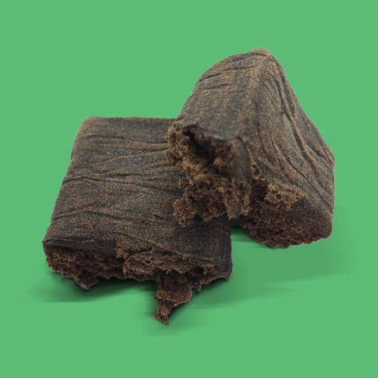 Two pieces of dark brown, crumbly CALI HASH AMNEZIA HAZE are displayed on a solid green background. Their soft, slightly fibrous texture features visible cracks and uneven edges, highlighting the product’s rich appearance.